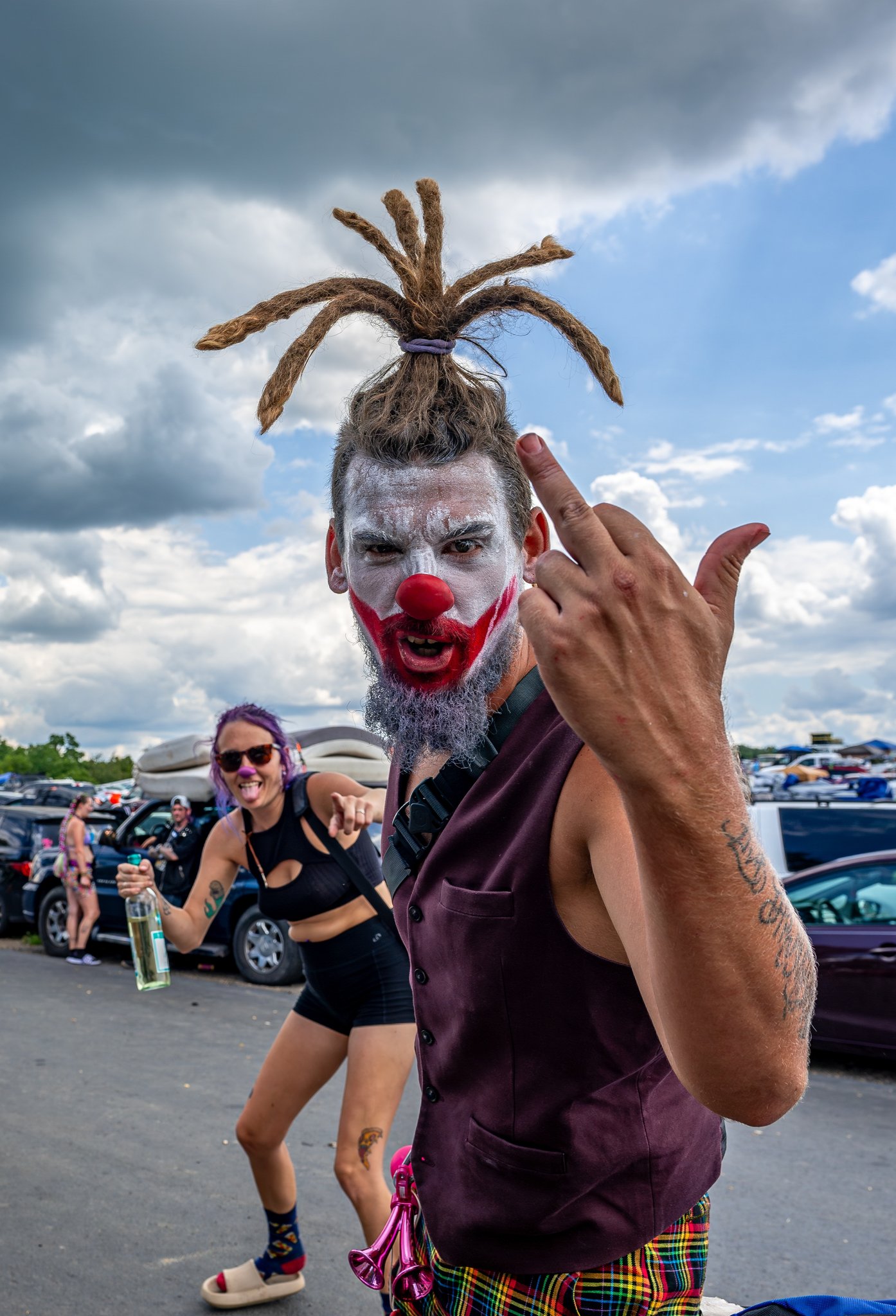 a juggalo with wild hair and red clown face makeup is showing his middle finger to the camera while a woman in the background points at the viewer and laughs. she is holding a bottle of vodka and the man's shorts are psychedelic..jpeg