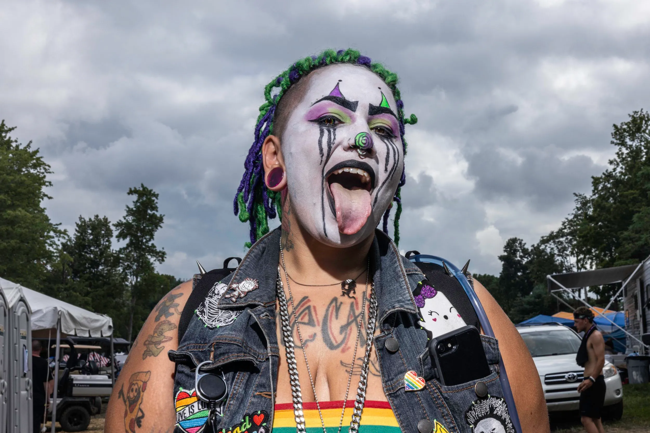 a sexy juggalo woman is sticking her tongue out and her cleavage is partly visible. she wears a denim vest cluttered with badges and pins.webp