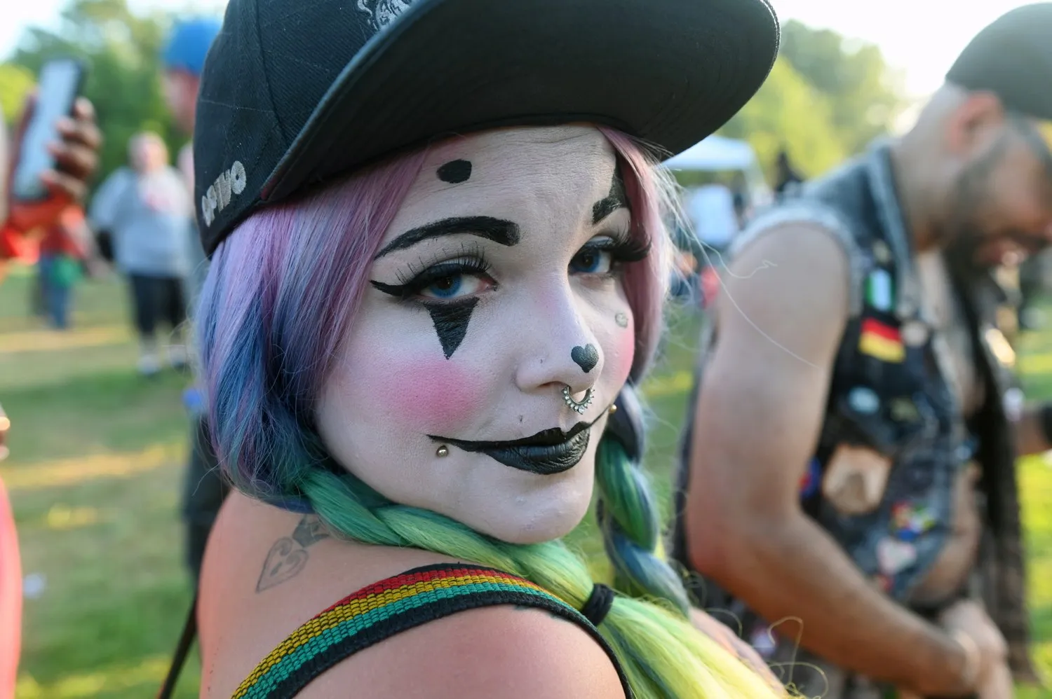 a juggalo is seen in a close-up photograph at a festival. she is wearing a baseball cap and purple hair. blurred in the background is a man in a vest, adorned with colourful badges, including the german flag.webp