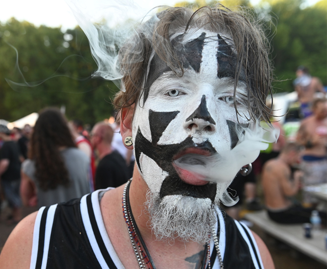 a juggalo with white eye contacts and black+white face paint is exhaling a vape cloud with wisps of smoke hanging around his head. his hair is greasy and stringy, and his beard has paint in it..jpg