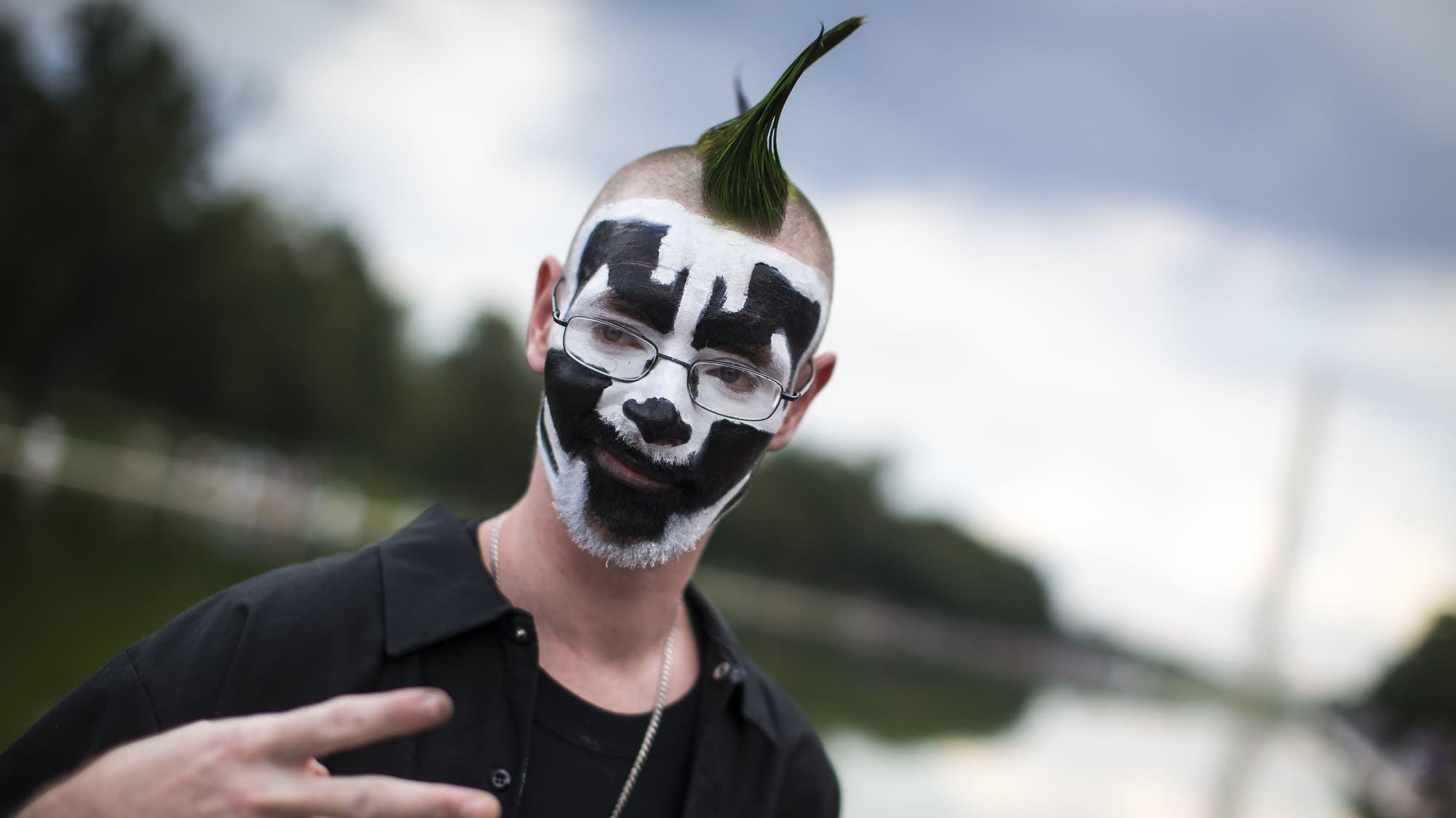 a nerdy ugly juggalo with painted facial hair and rectangular glasses throws up a peace sign. he has a green mowhawk and the background has bokeh.jpg