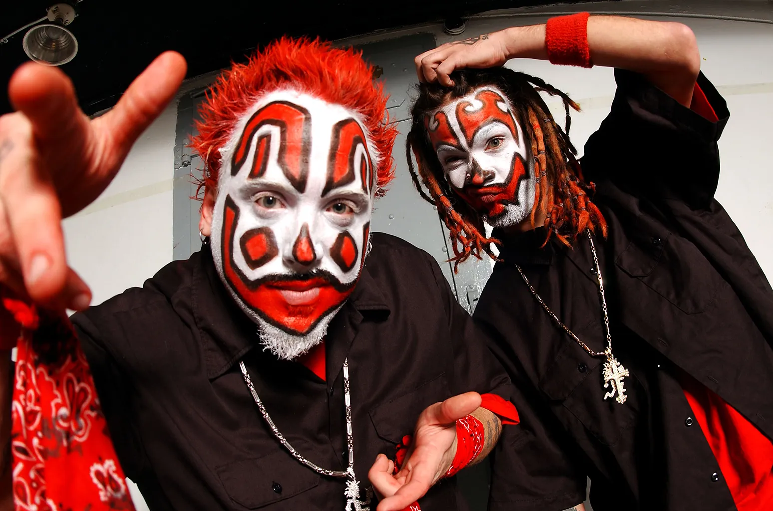 two juggalos seen with one making a hand jesture in red hair and makeup, while his friend with dreadlocks scratches his head and wears a black shirt and necklace.webp